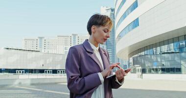 A beautiful young woman of European appearance with a short haircut, dressed in a business suit, walks in a modern metropolis smiling and using a tablet and looking around video