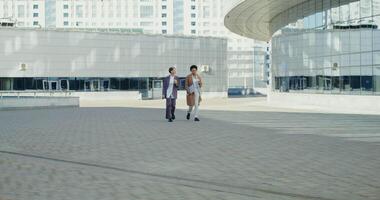 Two business women talking to each other and drinking coffee while walking through the business center of a modern city, on a sunny autumn day video