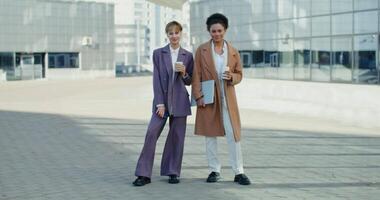 Two business women of different nationalities with glasses with coffee in their hands, are smiling looking at the camera, standing near the modern business center on a sunny autumn day video