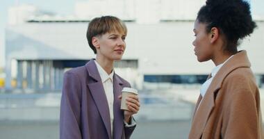 Two women in business suits are talking to each other while standing outdoors. A woman of European appearance drinks coffee from a disposable cup while talking with a colleague video