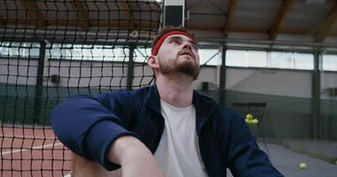 A young man of European appearance is resting after training, leaning his back and head on a tennis net stretched in the middle of the court, close-up video