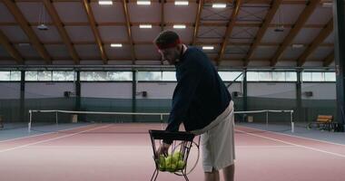 Tennis training alone without a partner, a man takes tennis balls from a basket full of balls and hits them with a racket, throwing them to the other side of the field. Filming from behind an athlete video