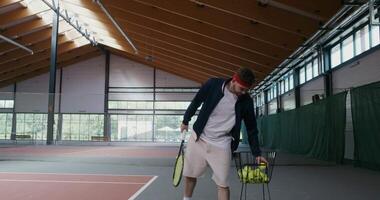 Tennis training alone without a partner, a young man takes tennis balls from a basket full of balls and hits them with a racket, throwing them to the other side of the field video