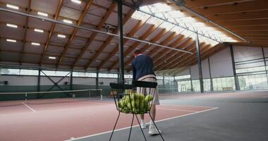 A young man of European appearance plays tennis alone, taking a tennis ball from a basket and tossing it with a racket over a net stretched in the middle of an indoor tennis court. Filming from behind video