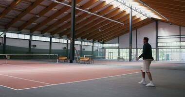 Young man and woman playing tennis on an indoor tennis court, bouncing the ball with a racket until the man loses a round by failing to throw the ball over the net stretched in the middle of the court video