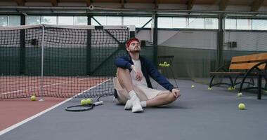A young man is resting after a workout with his back resting on a tennis net stretched in the middle of the court. There is a tennis racket nearby and lots of tennis balls scattered around video