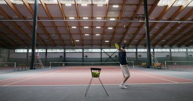 A young man of European appearance plays tennis alone, taking a tennis ball from a basket and tossing it with a racket over a net stretched in the middle of an indoor tennis court video