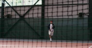 en ung man av europeisk utseende spelar stor tennis, slå de boll med en racket över och över om igen, studsande med varje racket träffa, filmning genom de netto, de teckning av de netto är synlig video