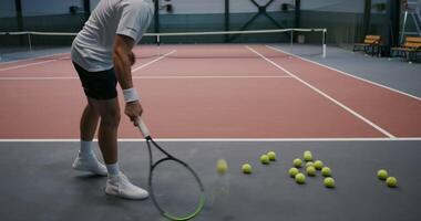 A young man of European appearance, dressed in a sports uniform, plays tennis alone, prepared many tennis balls on the floor and lifting them with racket strikes and throwing them over the net video