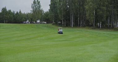 Panoramic view of the golf course with the golf car driving through it. Two men are about to play golf while moving from place to place in an electric car on a rainy day video