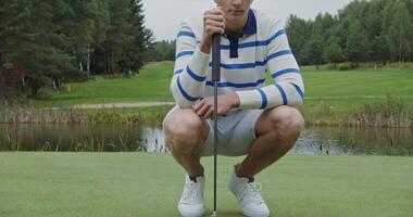 Young handsome man squatting in front of a golf ball assessing the distance to the hole, then hitting the ball with his club trying to roll it into the hole video