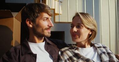The young family moves to a new home, lots of boxes and things are placed everywhere. A man and a woman are contentedly sitting on the couch and looking out the window, resting, close-up video