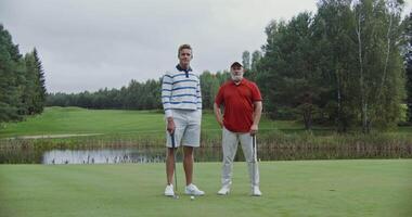 Two men of different ages are standing on the golf course with their legs crossed, and leaning on golf clubs and smiling while looking at the camera. video
