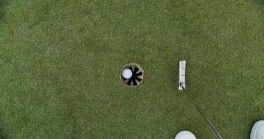 An overhead view of a golf course set on artificial turf. A man hits a ball with a golf club and it hits the target after spinning around the hole video