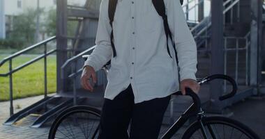A young man of European appearance in a knitted hat and with a backpack on his back is looking seriously at the camera, leaning on a bicycle video