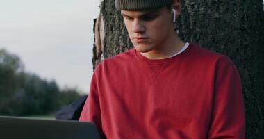 A young man of European appearance, in a knitted hat, uses a laptop, sitting on the ground under a tree in the open air. video