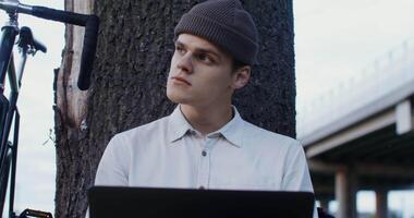 A young man of European appearance, in a knitted hat, uses a laptop, sitting on the ground under a tree in the open air. Bicycle Stands next to a man video