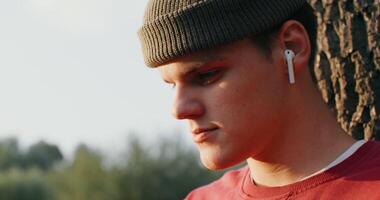 A young man of European appearance in a knitted hat and with wireless headphones in his ears sits on the ground under a tree and looks down intently, close-up, only his face is in the frame video