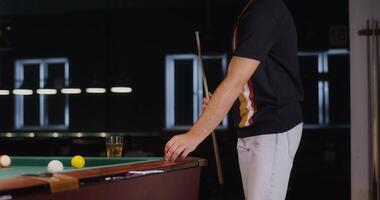 A young man of European appearance smears the tip of a cue with chalk and then hits the ball, bending over to the table, playing billiards. video