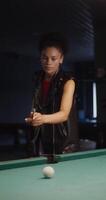 Beautiful young african american woman aiming intently and hitting a ball with a cue stick while playing billiards, vertical video