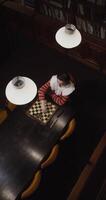Top view on a young woman of European appearance plays a chess game alone, thinking long concentratedly between moves, sitting at a long table in the library, vertical video