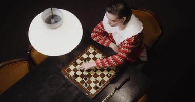 Top view on a young woman of European appearance plays a chess game alone, thinking long concentratedly between moves, sitting at a long table in the library, video