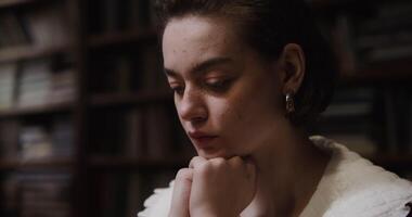 A young beautiful girl of European appearance with freckles and in large earrings is thinking intently, resting her chin with her hands and looking down at the table, close-up video