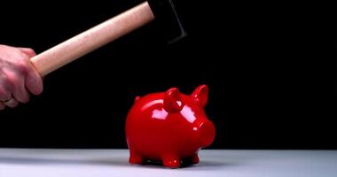 A man's hand smashes a red piggy bank in the shape of a pig with a hammer, fragments and coins fly in different directions, slow motion against a bright background video