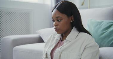Beautiful young african american woman in casual clothes with a serious look is typing on a laptop, while sitting on the floor in front of the sofa video