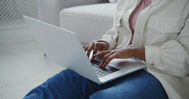 African american woman with red manicure typing on a laptop sitting on the floor in front of the sofa, close up video