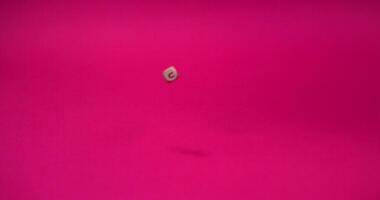 Brown beads in the shape of a square with letters written on them falling on a pink background, in slow motion video