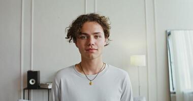 Handsome young curly-haired man of European appearance in casual clothes, with a pendant in the form of a fang smiles broadly, looking straight into the camera, laughing and rejoicing video