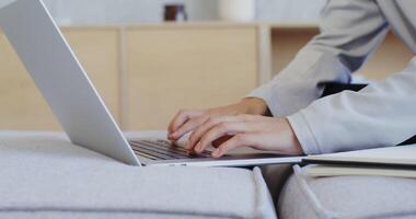 A woman of European appearance typing on a laptop and takes notes with a pen in a notebook sitting on a sofa in a lotus position, videography close-up video