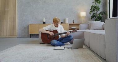 A man in casual clothes with beard looks at tutorial using laptop and plays the guitar while sitting on the floor at home, learning to use a musical instrument. Self-education in the modern world video