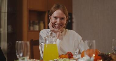 A beautiful red-haired young woman of European appearance with freckles smiling looks directly at the camera, sitting at the laid festive table video