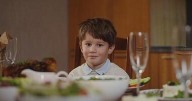 A dark-haired little boy of European appearance smiling looks directly at the camera, sitting at a laid festive table video