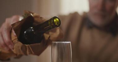 A gray-haired man with a beard pours wine from a bottle into a glass while sitting at a table, close-up shot video