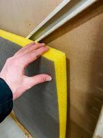 A person is gently touching and feeling the texture of a piece of foam that is affixed to a wall, possibly considering how it interacts with nearby surfaces like paint or wood stain photo