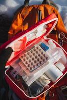 A person is holding a first aid kit in front of a colorful, sturdy backpack that is resting on the ground. The backpack appears to be made of durable material and is ideal for travel photo