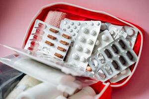 On a soft pink background, there is a vibrant red first aid kit that is generously filled with various pills, capsules, and bandages, ensuring preparedness for any medical need photo