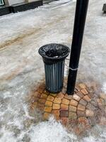 On a quiet, snowcovered sidewalk, a solitary trash can is placed next to a tall pole, which proudly stands amidst the serene winter landscape, adding a touch of urban charm photo
