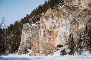 A significant and large rock formation that features a cave nestled within it is completely surrounded by a blanket of snow and tall trees, creating a picturesque winter landscape photo