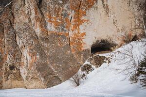There is a cave nestled in the side of a majestic mountain, which is beautifully surrounded by a blanket of pure white snow and rugged rocks that add to its stunning appearance photo