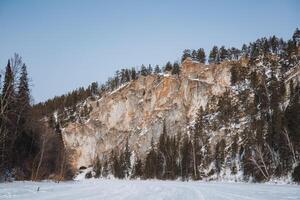 A stunning mountain that is entirely covered in a thick layer of white snow and tall trees, with a beautiful frozen lake visible prominently in the foreground of the scene photo