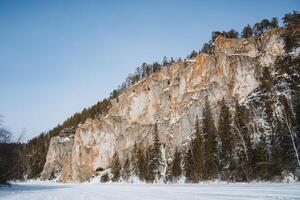 A substantial and imposing rocky cliff stands tall, beautifully surrounded by trees draped in thick, white snow, alongside a serene and frozen lake that reflects the winter landscape photo