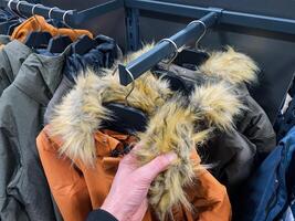 A person is currently holding a stylish jacket that features a luxurious fur hood while browsing in a clothing store, surrounded by various winter apparel and accessories photo