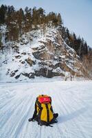 A vibrant yellow backpack is resting in the gleaming snow, positioned perfectly in front of a majestic, snowy mountain that towers in the background, creating a picturesque winter scene photo