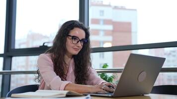 Beautiful woman working at computer and smiling. Female worker in a good mood works in an office. Workplace in big light office with large windows. video