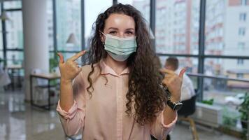 Dark-haired woman wearing mask in office. Girl showing her fingers at the mask, takes it off and smiles cheerfully. Happy lady holding a mask in her hand by the face. video