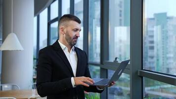 Male entrepreneur standing at the window and holding laptop in front of him. Business talk on the computer. Man talking and gesturing in front of notebook. video